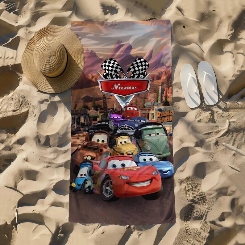 May include: A rectangular beach towel featuring characters from the animated movie "Cars." The towel has a brown background with images of race cars, the text "Name," and checkered flags. A straw hat and white flip-flops are on the sand.