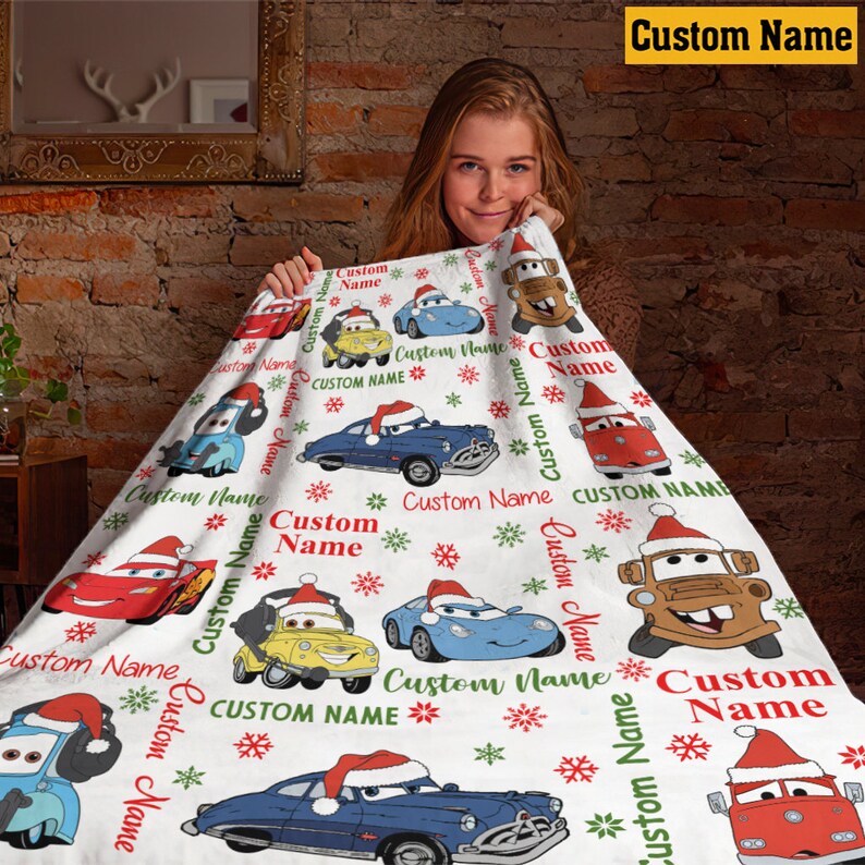 Puede incluir: Una manta de forro polar blanca con un patr&oacute;n repetitivo de coches de dibujos animados con gorros de Pap&aacute; Noel rojos y blancos, intercalados con copos de nieve rojos y verdes y el texto Custom Name en fuentes cursivas rojas y verdes.
