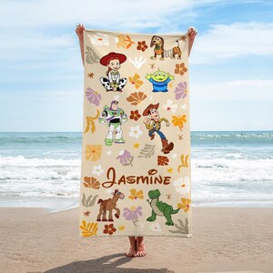 May include: A rectangular beach towel with a beige background featuring a repeating floral pattern in shades of orange, yellow, and purple. The towel displays Toy Story characters including Jessie, Slinky Dog, an alien, Buzz Lightyear, Woody, Bullseye, and Rex, with the name Jasmine printed in brown script.