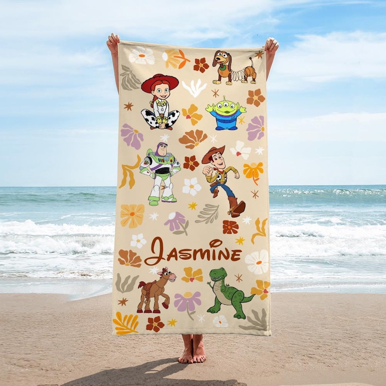 May include: A rectangular beach towel with a beige background featuring a repeating floral pattern in shades of orange, yellow, and purple. The towel displays Toy Story characters including Jessie, Slinky Dog, an alien, Buzz Lightyear, Woody, Bullseye, and Rex, with the name Jasmine printed in brown script.
