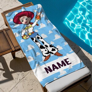 May include: A blue beach towel with a cartoon cowgirl character, red hat, and white clouds. The towel has a white border with the word "NAME" in dark purple letters. The towel is on a wooden sun lounger next to a swimming pool.