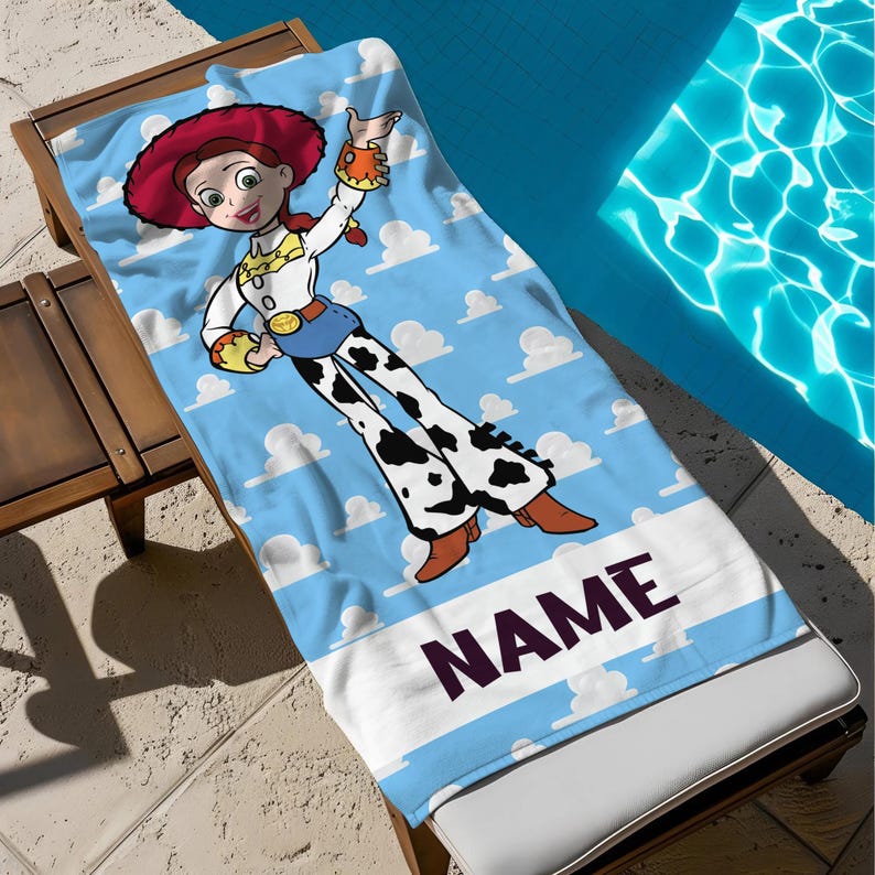 May include: A blue beach towel with a cartoon cowgirl character, red hat, and white clouds. The towel has a white border with the word "NAME" in dark purple letters. The towel is on a wooden sun lounger next to a swimming pool.