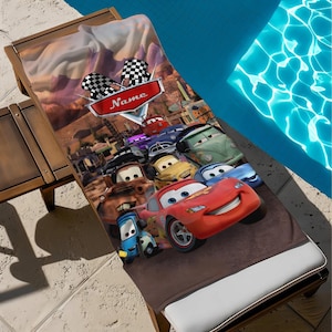 May include: A brown beach towel featuring a colorful Cars movie design. The towel showcases Lightning McQueen and other characters, with a checkered flag and the word "Name" on a red banner. The towel is placed on a wooden lounge chair near a pool.