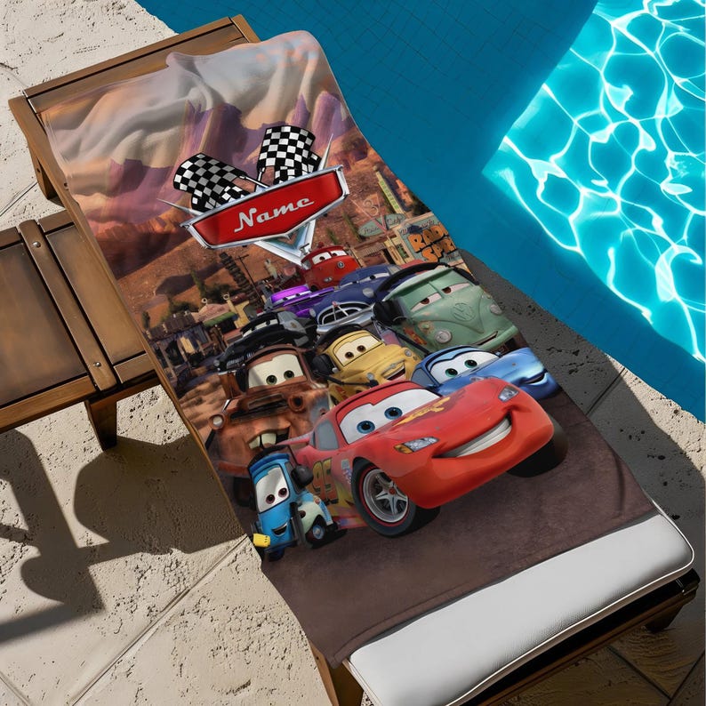 May include: A brown beach towel featuring a colorful Cars movie design. The towel showcases Lightning McQueen and other characters, with a checkered flag and the word "Name" on a red banner. The towel is placed on a wooden lounge chair near a pool.