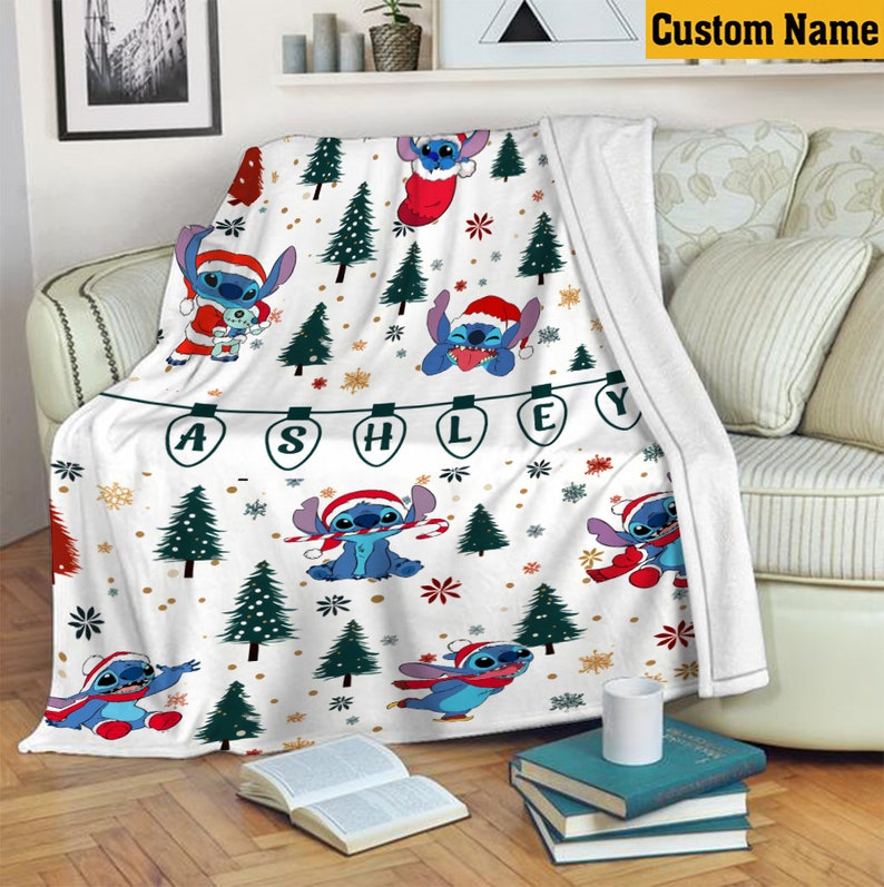 May include: A white fleece blanket featuring a repeating pattern of the character Stitch wearing a Santa hat, interspersed with green Christmas trees, snowflakes, and gold dots. A string of light bulbs spells out the name ASHLEY in black letters.