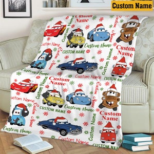Puede incluir: Una manta de forro polar blanca con un patr&oacute;n repetitivo de coches de dibujos animados con gorros de Pap&aacute; Noel, intercalados con copos de nieve rojos y verdes y el texto Custom Name impreso en rojo y verde sobre la tela.