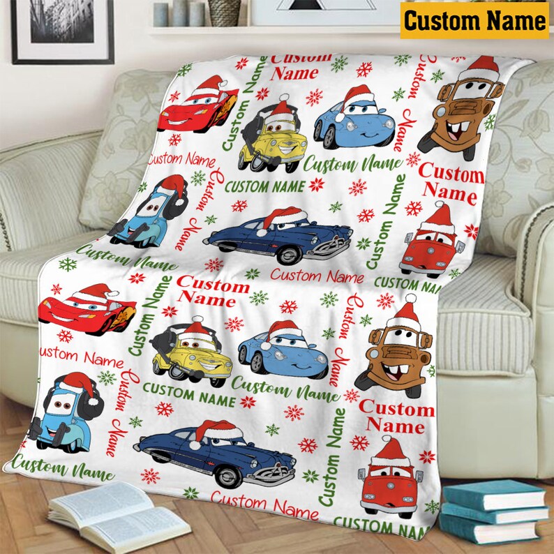 Puede incluir: Una manta de forro polar blanca con un patr&oacute;n repetitivo de coches de dibujos animados con gorros de Pap&aacute; Noel, intercalados con copos de nieve rojos y verdes y el texto Custom Name impreso en rojo y verde sobre la tela.