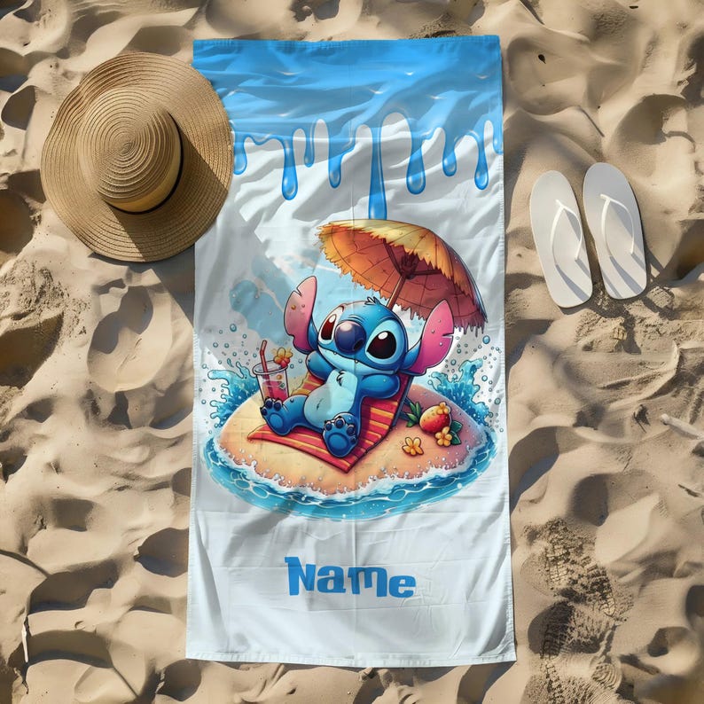 May include: A beach towel featuring a cartoon character relaxing on a tropical island with an umbrella. The towel has a blue and white design with the word "Name" printed on it. A straw hat and white flip-flops are next to the towel on a sandy surface.