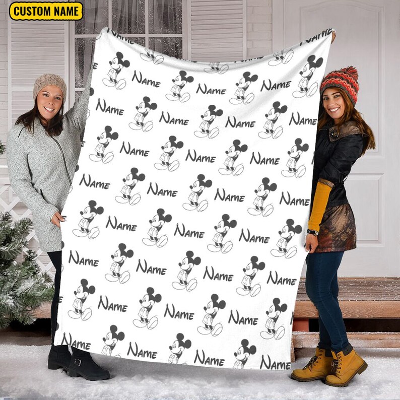 May include: A white fleece blanket featuring a repeating black and white pattern of Mickey Mouse figures and the word Name printed in a black serif font. The blanket includes a yellow rectangular label in the top left corner with the text CUSTOM NAME.
