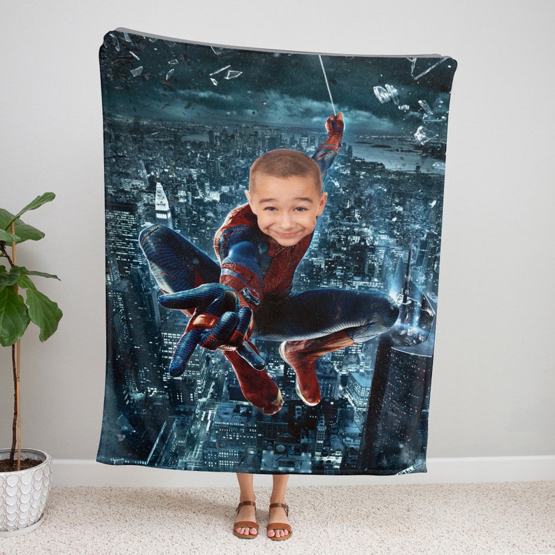 Puede incluir: Una manta de forro polar con la cara de un ni&ntilde;o superpuesta a un personaje de superh&eacute;roe balance&aacute;ndose por un horizonte urbano. El personaje lleva un traje rojo y azul con un dise&ntilde;o de telara&ntilde;a.