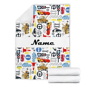 May include: A white fleece blanket featuring a repeating pattern of cartoon race cars, tow trucks, traffic lights, wrenches, steering wheels, and road signs. A horizontal white band across the centre displays the black text Name in script font.