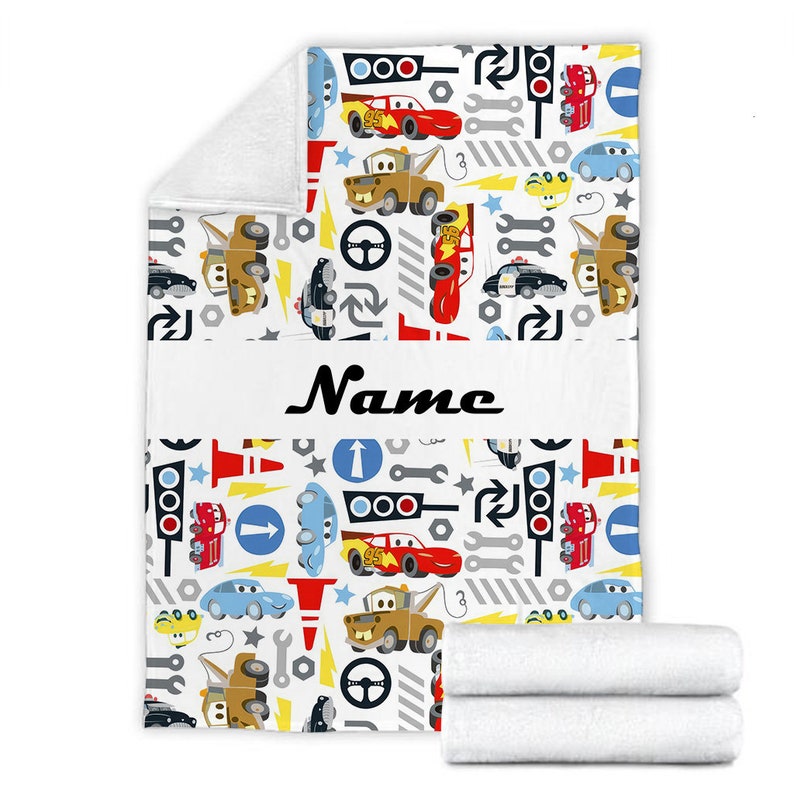 May include: A white fleece blanket featuring a repeating pattern of cartoon race cars, tow trucks, traffic lights, wrenches, steering wheels, and road signs. A horizontal white band across the centre displays the black text Name in script font.