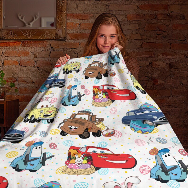 May include: A white fleece blanket with a colorful Easter-themed design featuring cartoon cars from the Disney Pixar movie Cars. The blanket features Lightning McQueen, Mater, and other characters from the movie. The blanket is decorated with Easter eggs, bunnies, and other Easter symbols.