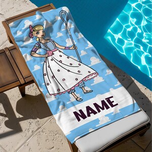 May include: A blue beach towel with a cartoon character in a white dress with pink details, holding a shepherd's crook. The towel has a sky and cloud design with the word "NAME" in a dark purple font.