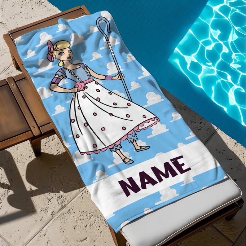 May include: A blue beach towel with a cartoon character in a white dress with pink details, holding a shepherd's crook. The towel has a sky and cloud design with the word "NAME" in a dark purple font.