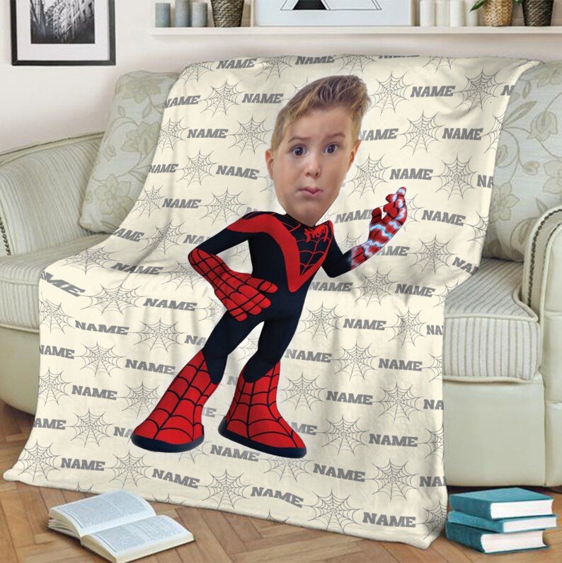 May include: A cream-coloured fleece blanket featuring a repeating pattern of grey spiderwebs and the word NAME printed in grey. Centered on the blanket is a cartoon-style character wearing a red and black superhero suit with a photo-realistic head.