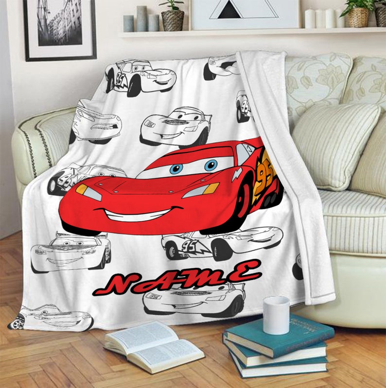 May include: A white fleece blanket featuring a repeating pattern of black and white line art of race cars, with one large, central, full-color red race car with blue eyes and the number 95, and the word NAME printed in red below the car.