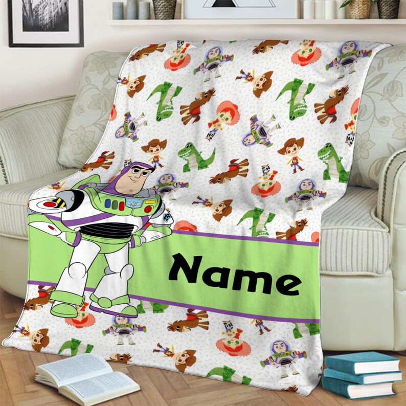 May include: A white fleece blanket featuring a repeating pattern of cartoon characters including a space ranger, a cowboy, a cowgirl, and a green dinosaur. A large space ranger figure stands on the left next to a green banner with the text Name.