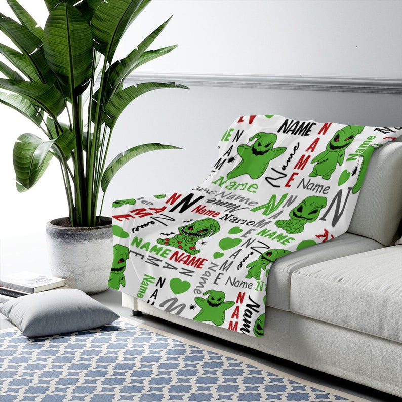 May include: White fleece blanket with a repeating pattern of green and red cartoon characters. The characters are a green monster with a stitched mouth and a red heart. The text "Name" is repeated throughout the pattern.