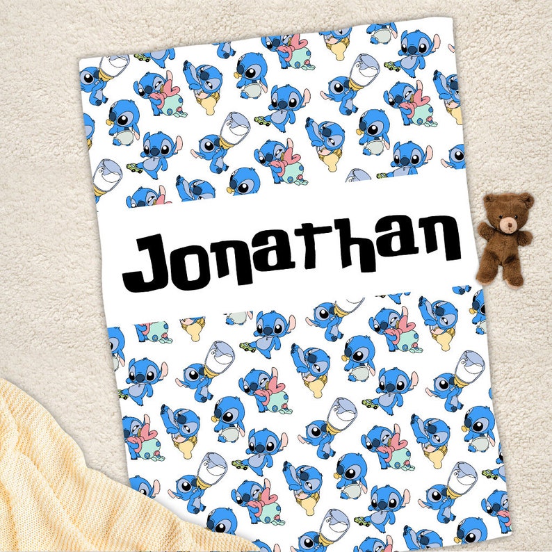 May include: A white blanket featuring a repeating pattern of blue alien characters holding baby bottles and wearing diapers, with the name Jonathan printed in large black letters across a central horizontal white band on the fabric.