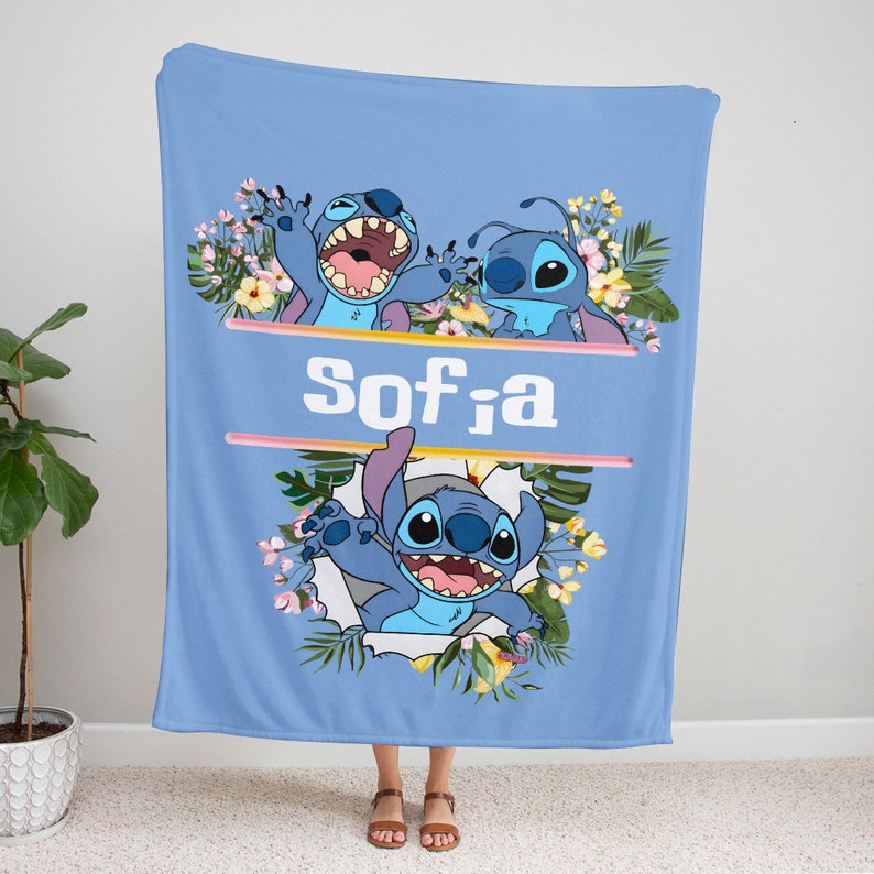 May include: A light blue fleece blanket featuring the character Stitch from Lilo and Stitch. The design includes tropical flowers, green leaves, and the name Sofia in white text between two horizontal lines. Stitch is shown in three poses.