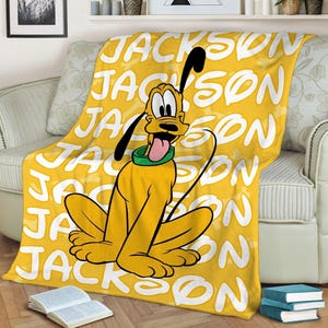 May include: Yellow fleece blanket with a cartoon image of Pluto the dog. The blanket has the name "Jackson" repeated in white text.