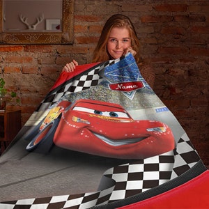 May include: A soft throw blanket featuring a large red race car character with blue eyes and a wide smile. The design includes a black and white checkered border, a stadium background, and a red shield-shaped emblem with the word Name printed in white.
