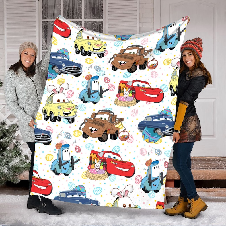 May include: A white fleece blanket with a repeating pattern of cartoon cars and Easter eggs. The cars are red, yellow, blue, and brown. The eggs are yellow, blue, pink, and green.