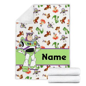 May include: A white fleece blanket featuring a repeating pattern of cartoon characters including a space ranger, a cowboy, a cowgirl, and a green dinosaur. A large space ranger figure stands on the left, next to a lime green banner with the text Name.