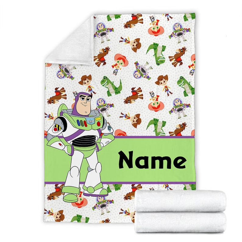 May include: A white fleece blanket featuring a repeating pattern of cartoon characters including a space ranger, a cowboy, a cowgirl, and a green dinosaur. A large space ranger figure stands on the left, next to a lime green banner with the text Name.