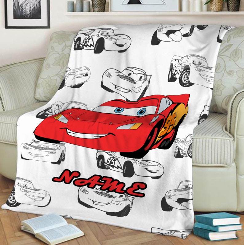 May include: A white fleece blanket featuring a repeating pattern of black and white line art race cars, with one large, central red race car with blue eyes and the number 95. The word NAME is printed in bold red letters below the central car.