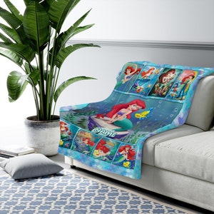 K&ouml;nnte beinhalten: Eine weiche blaue Wohndecke mit mehreren Szenen einer rothaarigen Meerjungfrau und eines gelben Fisches. Das Design zeigt eine gro&szlig;e zentrale Illustration der Meerjungfrau auf einem Felsen sitzend mit dem Wort NAME darunter gedruckt.