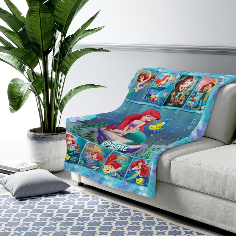 K&ouml;nnte beinhalten: Eine weiche blaue Wohndecke mit mehreren Szenen einer rothaarigen Meerjungfrau und eines gelben Fisches. Das Design zeigt eine gro&szlig;e zentrale Illustration der Meerjungfrau auf einem Felsen sitzend mit dem Wort NAME darunter gedruckt.