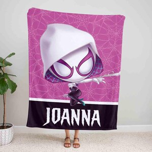 K&ouml;nnte beinhalten: Eine rosa und schwarze Fleece-Decke mit einem Cartoon-Bild einer wei&szlig;en Spinnenfrau in einem schwarzen Anzug mit einem Spinnennetz-Design. Die Decke tr&auml;gt den Namen "JOANNA" unten aufgedruckt.