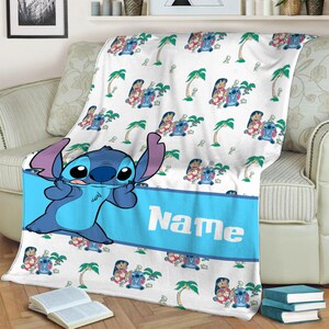 K&ouml;nnte beinhalten: Eine wei&szlig;e Fleecedecke mit einem sich wiederholenden Muster aus Lilo und Stitch-Figuren und Palmen. Ein breites blaues horizontales Band in der Mitte zeigt das Wort Name in wei&szlig;en Buchstaben, mit einer gro&szlig;en Stitch-Figur auf der linken Seite.