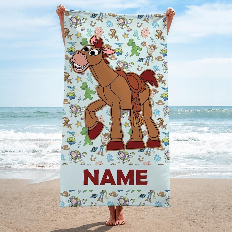 May include: A light blue beach towel featuring a large brown horse character with a saddle in the center. The background has a repeating pattern of cartoon characters, stars, and cacti. The word NAME is printed in large dark red letters at the bottom.