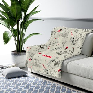 Peut inclure: Un plaid de couleur cr&egrave;me orn&eacute; d'un motif r&eacute;p&eacute;titif en traits noirs repr&eacute;sentant divers personnages de dessins anim&eacute;s, dont un ranger de l'espace, un cow-boy et un dinosaure. Le design inclut des accents rouges, des &eacute;toiles et le mot NAME en rouge.