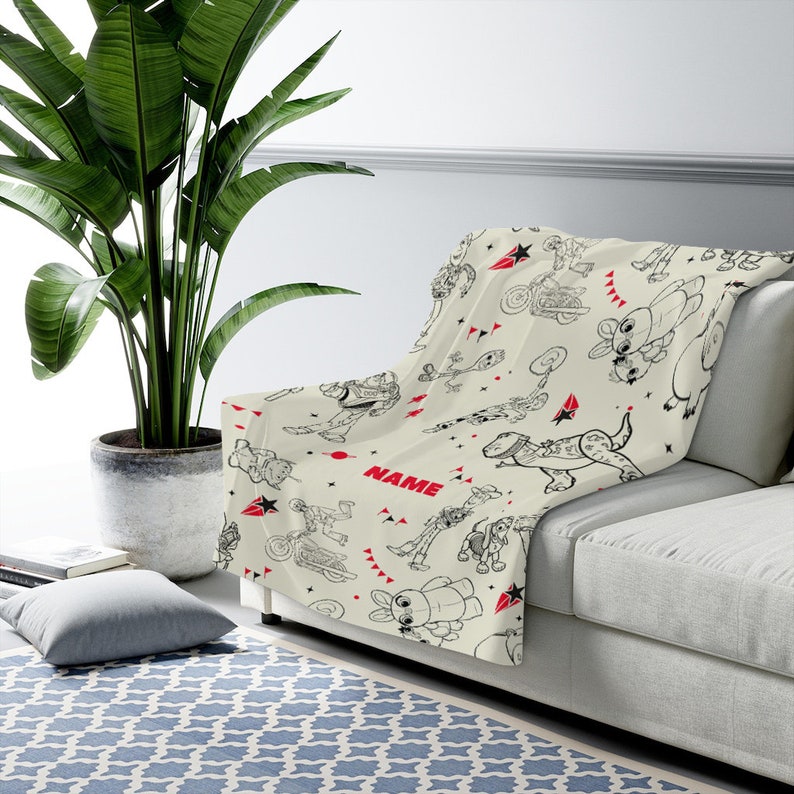 Peut inclure: Un plaid de couleur cr&egrave;me orn&eacute; d'un motif r&eacute;p&eacute;titif en traits noirs repr&eacute;sentant divers personnages de dessins anim&eacute;s, dont un ranger de l'espace, un cow-boy et un dinosaure. Le design inclut des accents rouges, des &eacute;toiles et le mot NAME en rouge.