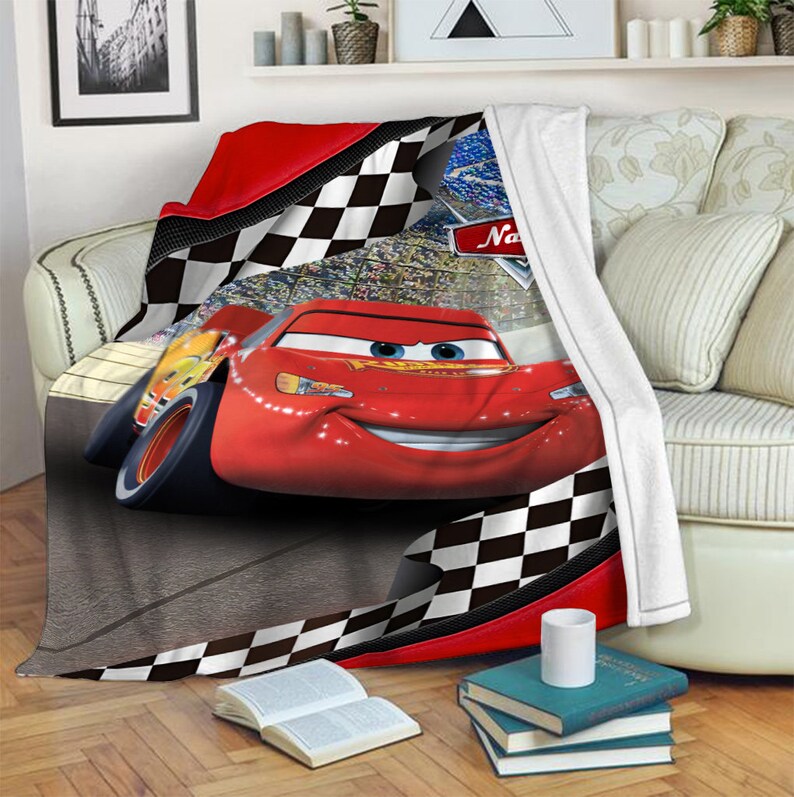 May include: A plush throw blanket featuring a red race car with blue eyes and a wide smile on a racetrack. The design includes a black and white checkered border, a stadium crowd background, and a red logo with the text Na in the upper corner.