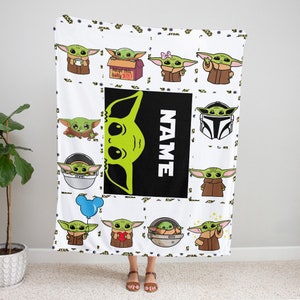 May include: A white fleece blanket featuring a repeating pattern of green alien characters in brown robes. A central black rectangular panel displays a large green alien head graphic next to the white vertical text NAME and a box labeled Adopt this baby JEDI.