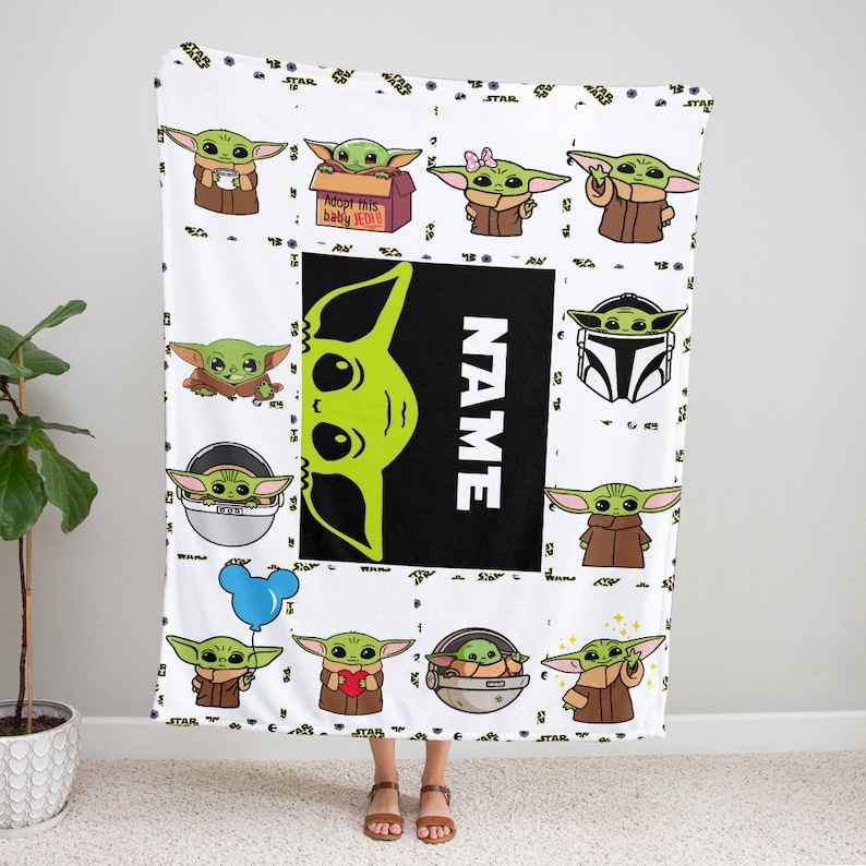 May include: A white fleece blanket featuring a repeating pattern of green alien characters in brown robes. A central black rectangular panel displays a large green alien head graphic next to the white vertical text NAME and a box labeled Adopt this baby JEDI.