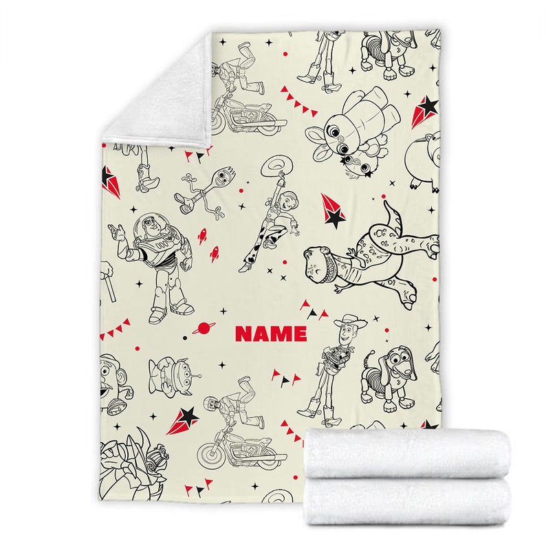 Peut inclure: Une couverture en polaire cr&egrave;me avec un motif r&eacute;p&eacute;titif en traits noirs repr&eacute;sentant divers personnages anim&eacute;s, dont un ranger de l'espace, un cow-boy et un dinosaure. Le design inclut des accents rouges, des &eacute;toiles et le mot NAME en rouge.