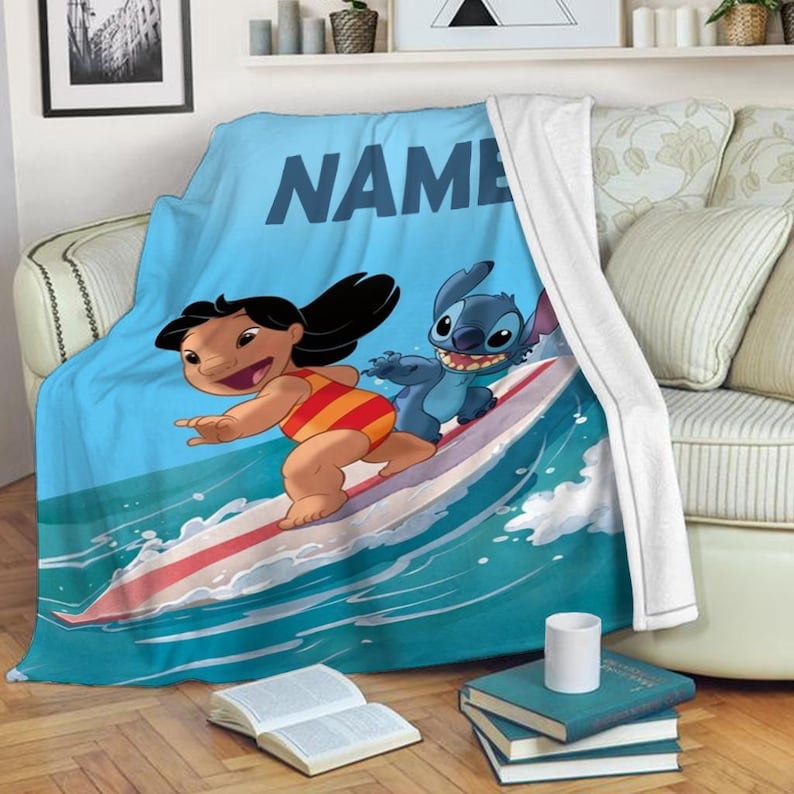 Peut inclure: Une couverture en polaire bleue avec un nom personnalis&eacute;, "NAME", et une image de dessin anim&eacute; de Lilo et Stitch sur une planche de surf sur une vague.