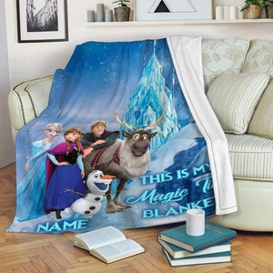 May include: A blue fleece throw blanket featuring a printed graphic of Elsa, Anna, Kristoff, Sven, and Olaf in front of an ice castle. The text reads THIS IS MY Magic TIME BLANKET and NAME. The blanket has a white underside and a soft texture.