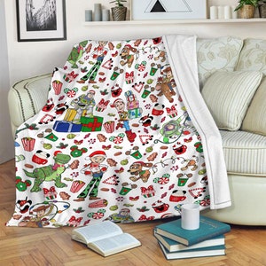 Puede incluir: Una manta de forro polar blanca con un patr&oacute;n navide&ntilde;o repetitivo de personajes de Toy Story como Woody, Buzz Lightyear, Jessie, Rex y Slinky Dog, intercalados con iconos festivos como regalos, bastones de caramelo y tazas de cacao.