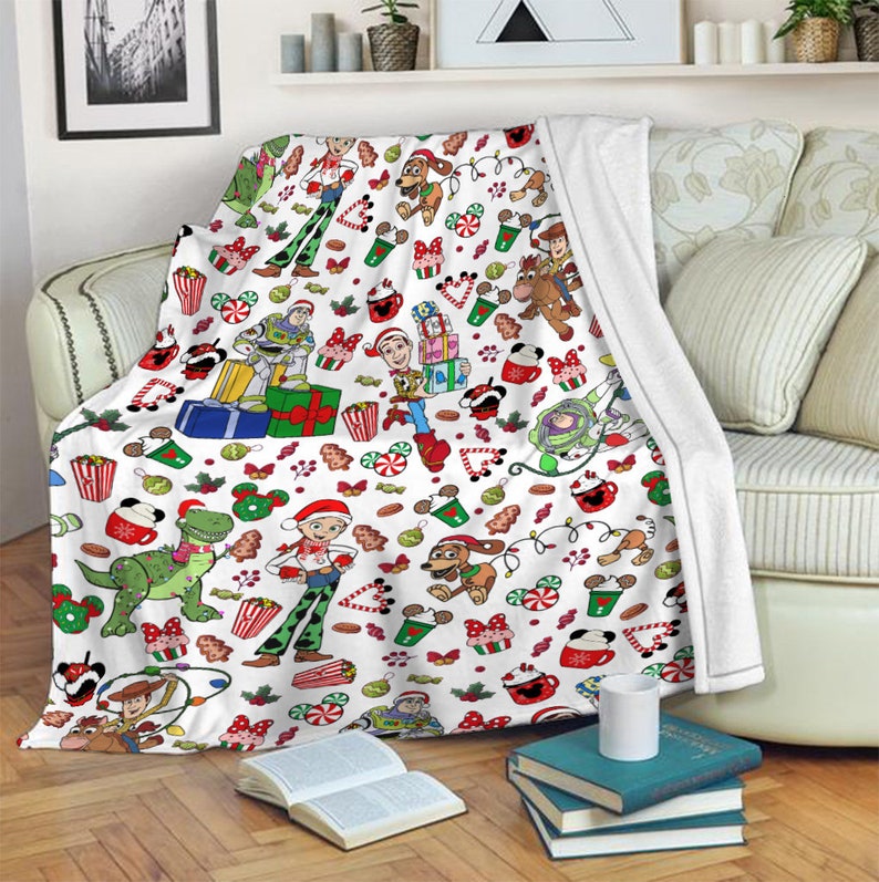 Puede incluir: Una manta de forro polar blanca con un patr&oacute;n navide&ntilde;o repetitivo de personajes de Toy Story como Woody, Buzz Lightyear, Jessie, Rex y Slinky Dog, intercalados con iconos festivos como regalos, bastones de caramelo y tazas de cacao.