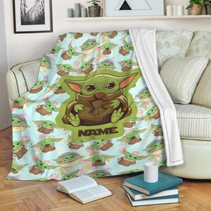 Może przedstawiać: Miękki, niebieski i zielony koc polarowy z powtarzającym się wzorem postaci z kresk&oacute;wek. Postać jest zielona i ma duże oczy i uszy. Koc ma nadrukowane słowo "NAME".