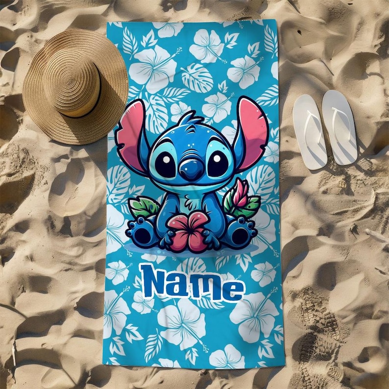 May include: A blue beach towel featuring a cartoon character with large ears, holding a flower. The towel has a floral pattern and the word "Name" in white. A straw hat and white flip-flops are on the sand.