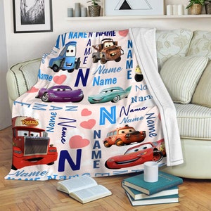 May include: A white fleece blanket featuring a repeating pattern of characters from the movie Cars, including a red truck, a purple car, a teal car, a brown tow truck, and a red race car, interspersed with pink hearts and the word Name.