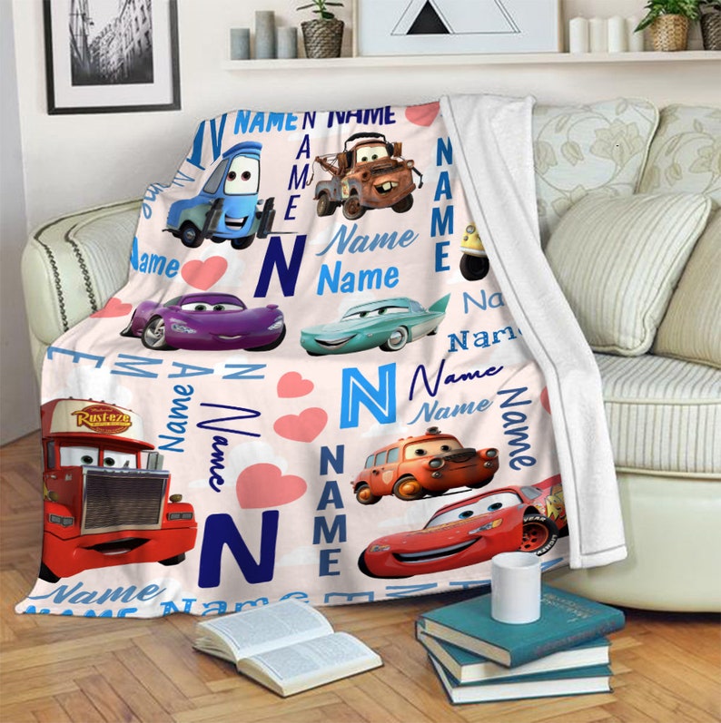 May include: A white fleece blanket featuring a repeating pattern of characters from the movie Cars, including a red truck, a purple car, a teal car, a brown tow truck, and a red race car, interspersed with pink hearts and the word Name.